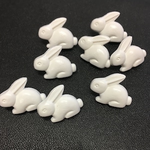 May include: A collection of eight small, white bunny figurines. Each bunny is in a seated position with long ears and a rounded body. The bunnies are arranged on a dark surface, creating a charming and whimsical display.
