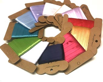 May include: A collection of 10 spools of satin ribbon in various colors, including white, pink, blue, green, and red. Each spool is wrapped in brown paper with a label that says "Beautiful moments".