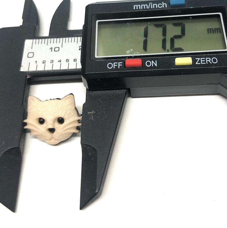 Pu&ograve; includere: Un primo piano di un bottone a forma di viso di gatto beige con occhi neri e baffi. Il bottone viene misurato con un calibro, che indica 17,2 mm.