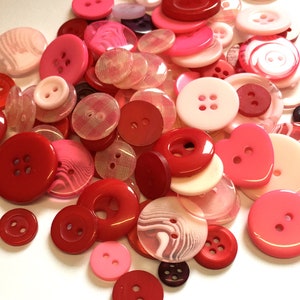 Puede incluir: Una colección de botones rojos y rosas de varios tamaños y formas. Los botones incluyen diseños redondos, ovalados y en forma de corazón, con estilos de dos y cuatro agujeros. Algunos botones son translúcidos, otros opacos.