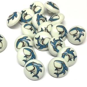 May include: A collection of white ceramic buttons with blue shark illustrations. The buttons are round and have a two-hole design.