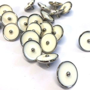 May include: A pile of white and silver round buttons with a raised centre. The buttons have a silver rim and a white centre with a small silver dot in the middle.