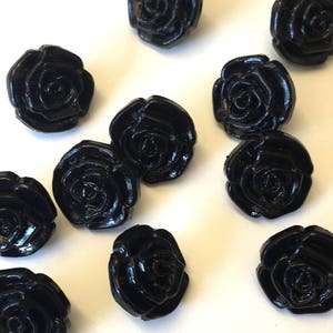 Könnte beinhalten: Eine Sammlung glänzender, schwarzer Rosenknöpfe. Die Knöpfe sind gleichmäßig verteilt und haben ein detailliertes Blumendesign. Diese dekorativen Knöpfe eignen sich ideal zum Nähen, Basteln oder um Kleidungsstücken eine einzigartige Note zu verleihen.
