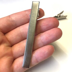 Puede incluir: Una pinza para el pelo de metal plateado, de forma rectangular, con un cierre de bisagra. La pinza se sostiene en una mano sobre un fondo blanco. Otra pinza es visible en el fondo.
