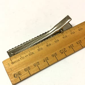 Puede incluir: Una pinza para el pelo de metal plateado con un dise&ntilde;o texturizado y acanalado. La pinza se mide con una regla de madera, mostrando una longitud de aproximadamente 7,6 cm.