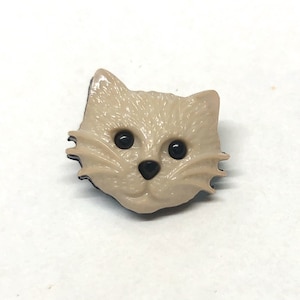 Pu&ograve; includere: Un bottone di plastica beige con un design a forma di gatto. Il gatto ha occhi neri, un naso nero e un contorno nero.