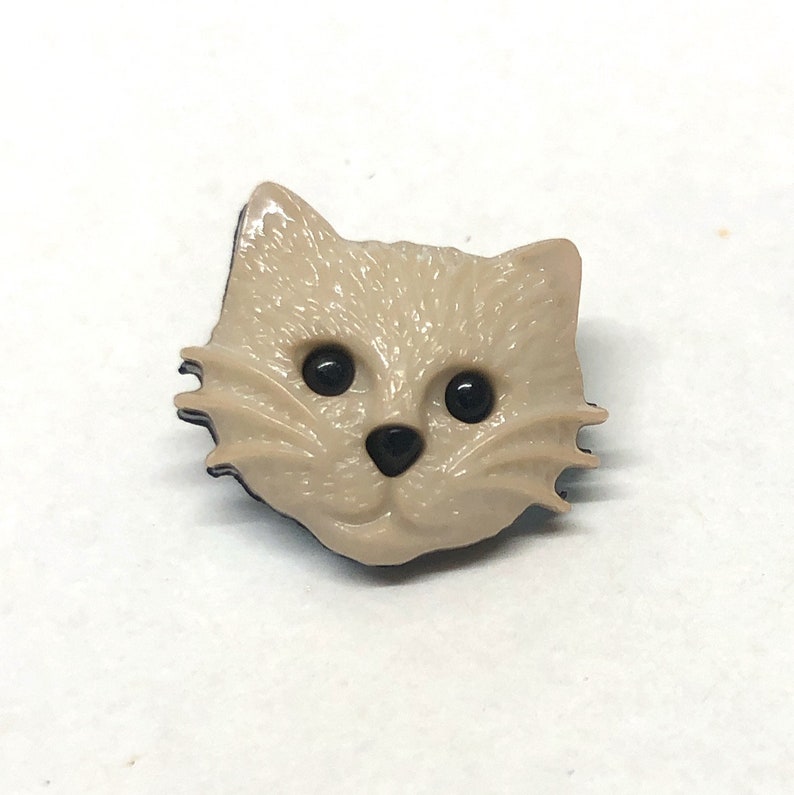 Pu&ograve; includere: Un bottone di plastica beige con un design a forma di gatto. Il gatto ha occhi neri, un naso nero e un contorno nero.