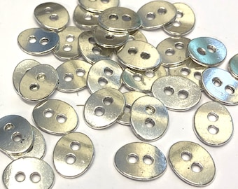 May include: A collection of silver-colored, oval-shaped buttons with two holes each. The buttons are scattered across a white surface, reflecting light and appearing slightly tarnished. These buttons are suitable for sewing and craft projects.