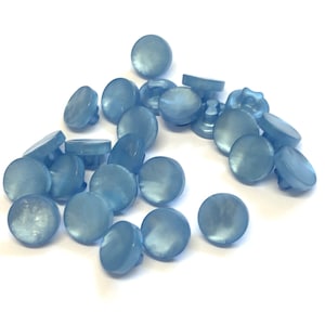 May include: A pile of light blue, pearlescent buttons. The buttons are round with a domed top and a shank on the underside. They are scattered on a white background. These buttons are suitable for various sewing and craft applications.
