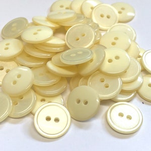 Może przedstawiać: Stosik kremowych plastikowych guzików z dwoma otworami każdy. Guziki są gładkie i błyszczące.