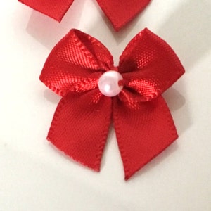 以下が含まれることがあります： 鮮やかな赤色のサテンリボンの小さなリボンがいくつか写っています。各リボンの中心には、小さな薄いピンク色のパールが付いています。リボンは白い面に配置されています。これらのリボンは、工芸品、ギフト、または装飾に適しています。