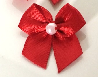 May include: Close-up of several small, bright red satin bows. Each bow has a small, light pink pearl in the center. The bows are arranged on a white surface. These bows are suitable for crafts, gifts, or decorations.