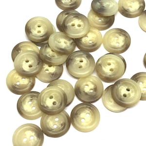 Può includere: Una collezione di 20 bottoni in plastica beige e marrone con due fori ciascuno. I bottoni hanno un motivo screziato e un bordo leggermente rialzato.