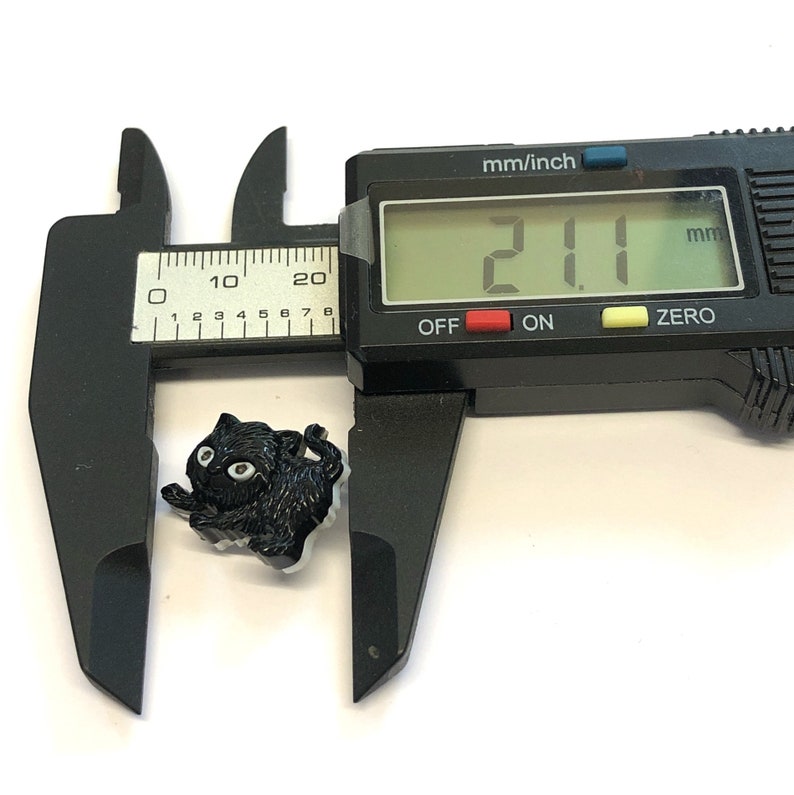 May include: A black cat button with white eyes, measuring 21.0 mm in diameter, as shown on a digital caliper.