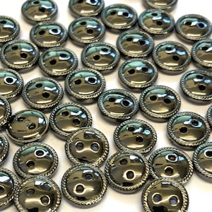 Op de afbeelding: Een verzameling zwarte metalen knopen met een verhoogde, touwachtige rand. De knopen hebben twee gaten en een glanzende, reflecterende afwerking.