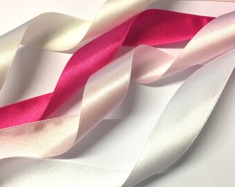 Könnte beinhalten: Drei Satinbänder in Weiß, Elfenbein und Rosa. Die Bänder sind kreuzweise angeordnet und zeigen ihre glatte Textur und leuchtenden Farben.