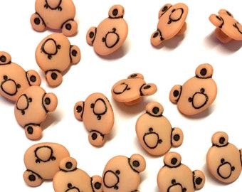 May include: A collection of 15 peach-colored plastic buttons with a brown bear face design.