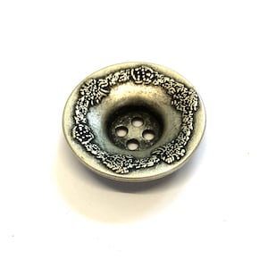 Pode incluir: Um botão prateado com um design floral decorativo ao redor da borda. O botão tem quatro furos no centro.