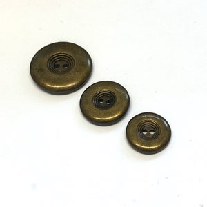 May include: Three round, brass-coloured buttons with two holes each. The buttons are different sizes, with the largest button at the top of the image and the smallest button at the bottom.