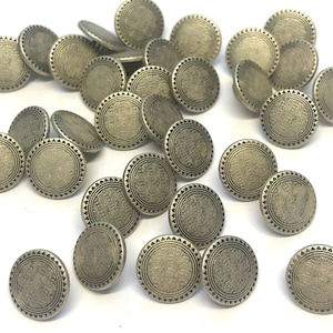 Könnte beinhalten: Eine Sammlung von 20 antiken silberfarbenen Knöpfen mit einem dekorativen, geprägten Design. Jeder Knopf ist rund mit einer erhabenen, strukturierten Mitte und einem gewellten Rand.