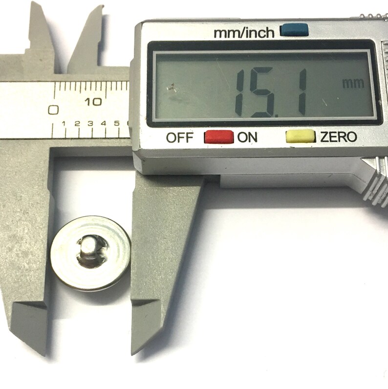 May include: A silver metal button with a diameter of 15.1 millimeters, measured with a digital caliper.