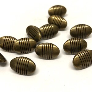 May include: A collection of oval-shaped, antique brass buttons with vertical, parallel lines etched into their surface. The buttons are scattered on a white background, showcasing their metallic sheen and detailed design.