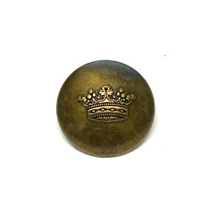 Pode incluir: Um botão de metal dourado com um design de coroa. A coroa tem um design detalhado com uma superfície texturizada.