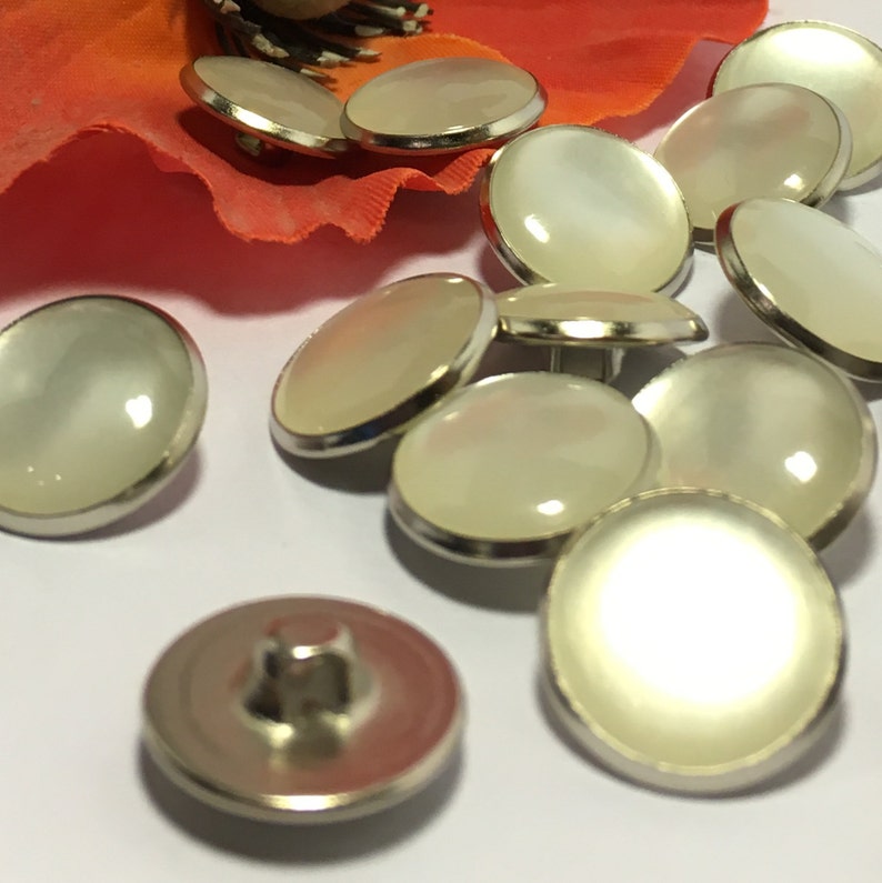 May include: A group of 10 white pearl buttons with silver metal rims. The buttons are arranged in a semi-circle on a white surface.