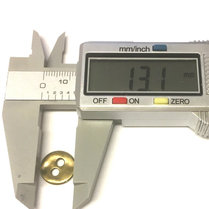 May include: A brass button with two holes is measured with a digital caliper. The measurement reads 13.1 mm, which is approximately 0.5 inches. The button is round and has a slightly raised edge.