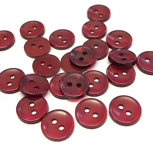 May include: A group of 18 red plastic buttons with two holes each. The buttons are scattered on a white surface.