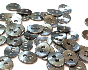 May include: A collection of silver-colored, oval-shaped buttons with two holes each. The buttons are scattered across a white surface, with varying angles and slight reflections. These buttons are suitable for sewing and craft projects.