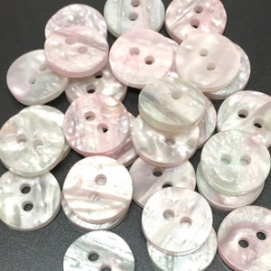Puede incluir: Una colección de botones de concha iridiscente rosa y blanco de 2 agujeros. Los botones son redondos y tienen un acabado liso y pulido.