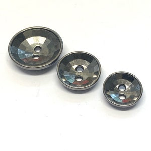 Pode incluir: Três botões redondos pretos de dois furos com um design facetado. Os botões estão dispostos em ordem de tamanho, com o botão maior à esquerda e o botão menor à direita.