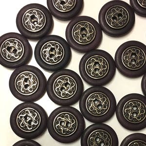 Può includere: Una collezione di 15 bottoni rotondi di colore viola e argento con un design decorativo. I bottoni hanno due fori.