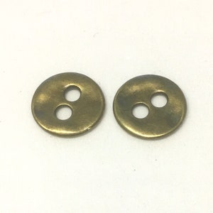 May include: Two antique brass-coloured buttons, each with two holes. The buttons are round and have a slightly aged appearance. They are set against a white background.