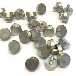 May include: A collection of silver metal buttons with a flat, round top and a small, raised post on the bottom. The buttons are scattered on a white surface.