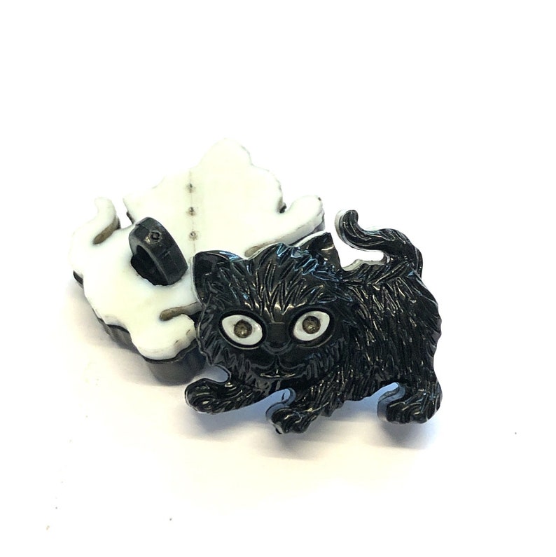 May include: Black plastic button with a kitten design. The kitten has white eyes and a black body.