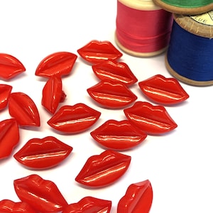 May include: A collection of glossy, red lip-shaped buttons scattered on a white surface. The buttons are a vibrant shade of red, with a smooth, reflective finish. Spools of thread in red, green, and blue are in the background.
