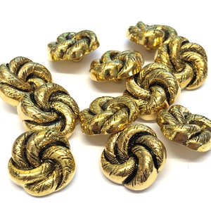 May include: A collection of gold-colored buttons with a textured, knotted design. The buttons are round with a detailed, swirling pattern. The buttons are likely for sewing or crafting projects.