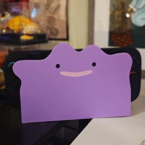 Könnte beinhalten: Ein lila Zubehör in Ditto-Form mit schwarzen Augen und einem rosa Mund, das für eine schwarze Spielkonsole konzipiert wurde. Das Zubehör besteht aus einem weichen Material und dient zum Schutz der Konsole.