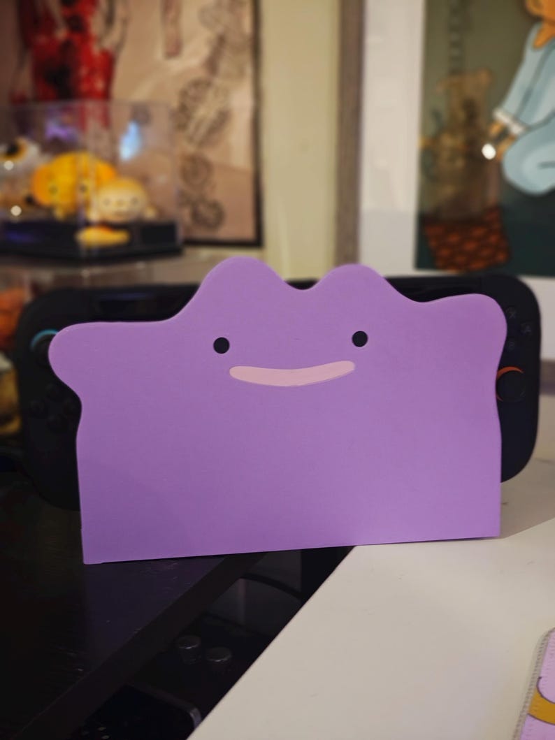 K&ouml;nnte beinhalten: Ein lila Zubeh&ouml;r in Ditto-Form mit schwarzen Augen und einem rosa Mund, das f&uuml;r eine schwarze Spielkonsole konzipiert wurde. Das Zubeh&ouml;r besteht aus einem weichen Material und dient zum Schutz der Konsole.