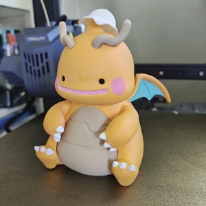 May include: A 3D printed orange dragon figurine with a tan belly, small white claws, and a white bucket hat. The dragon has pink circular cheeks, a pink mouth, two tan antennae, and small blue wings. The surface shows visible 3D print layer lines.