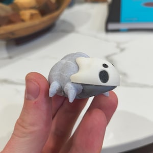 Peut inclure: Petite figurine grise et blanche représentant une créature avec un corps arrondi et de petites pattes. La figurine a une tête blanche avec des taches noires et est tenue dans une main.