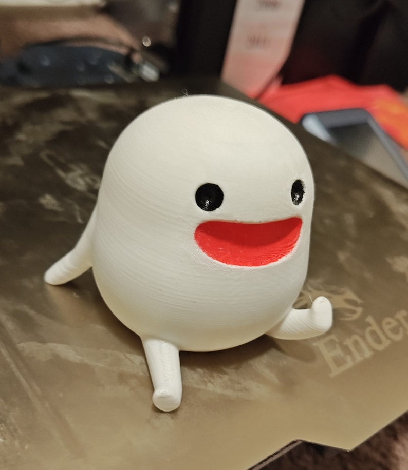 May include: White 3D printed figurine of a smiling ghost with red lips and black eyes. The ghost has two small arms and two long legs.