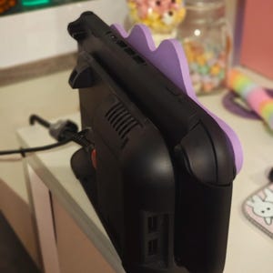 K&ouml;nnte beinhalten: Schwarze Ladestation mit lila, wellenf&ouml;rmiger Halterung f&uuml;r eine tragbare Spielkonsole. Das Dock hat ein schlankes Design mit L&uuml;ftungsschlitzen und Anschl&uuml;ssen. Ein schwarzes Netzkabel ist eingesteckt.