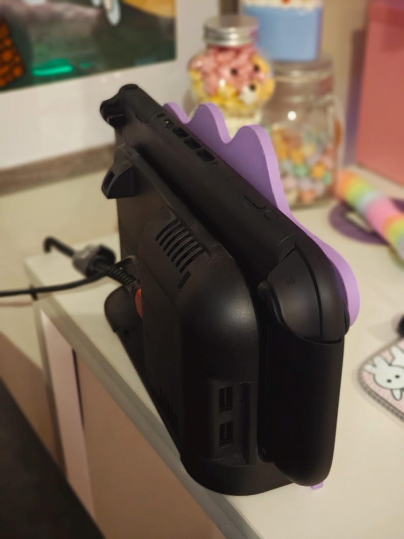 K&ouml;nnte beinhalten: Schwarze Ladestation mit lila, wellenf&ouml;rmiger Halterung f&uuml;r eine tragbare Spielkonsole. Das Dock hat ein schlankes Design mit L&uuml;ftungsschlitzen und Anschl&uuml;ssen. Ein schwarzes Netzkabel ist eingesteckt.