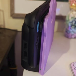 K&ouml;nnte beinhalten: Eine schwarze Spielkonsole mit einem hellvioletten, katzenf&ouml;rmigen Zubeh&ouml;rteil. Die Konsole hat blaue Kn&ouml;pfe und ein schlankes Design. Das Zubeh&ouml;r ist einfarbig und verleiht dem Ger&auml;t ein dekoratives Element.