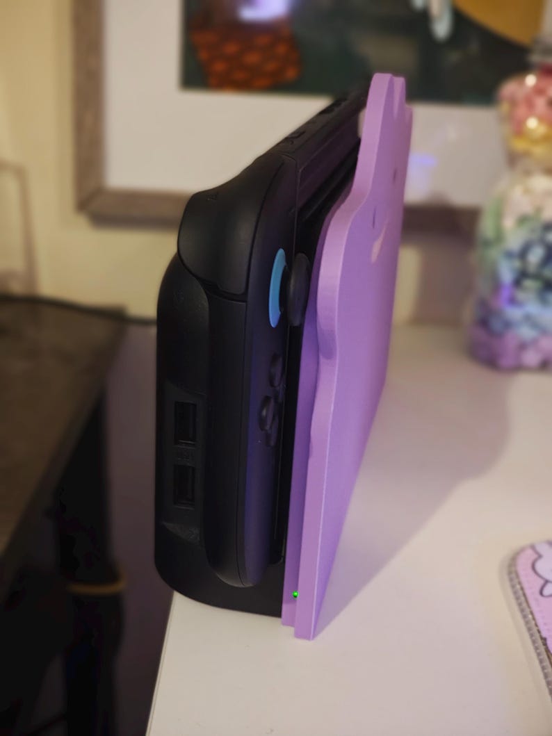 K&ouml;nnte beinhalten: Eine schwarze Spielkonsole mit einem hellvioletten, katzenf&ouml;rmigen Zubeh&ouml;rteil. Die Konsole hat blaue Kn&ouml;pfe und ein schlankes Design. Das Zubeh&ouml;r ist einfarbig und verleiht dem Ger&auml;t ein dekoratives Element.