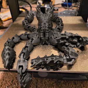 Può includere: Un polpo articolato stampato in 3D di colore nero con un design dettagliato e spinoso. Il polpo è posizionato su una superficie grigia, con i tentacoli distesi.
