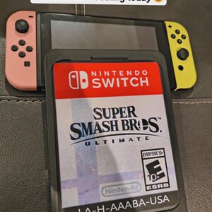 Könnte beinhalten: Eine Nintendo Switch-Konsole mit rosa und gelben Joy-Cons liegt über einer Spielkassettenhülle. Auf der Hülle sind der Spieletitel "Super Smash Bros. Ultimate" und das Nintendo Switch-Logo zu sehen. Der Text "LA-H-AAABA-USA" ist sichtbar.